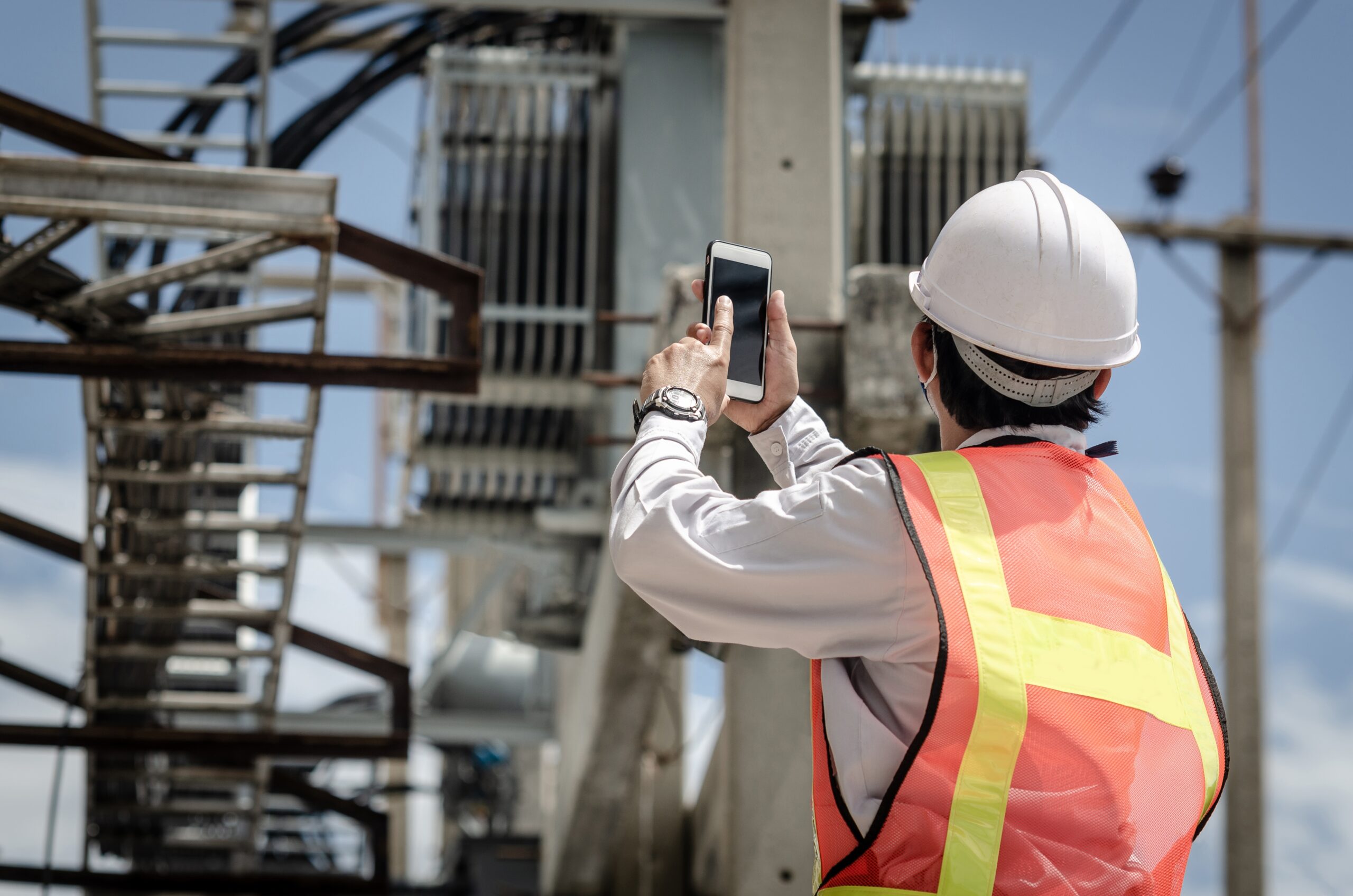 Electrician,Holds,A,Smartphone,Against,The,Background,Of,A,Transformer